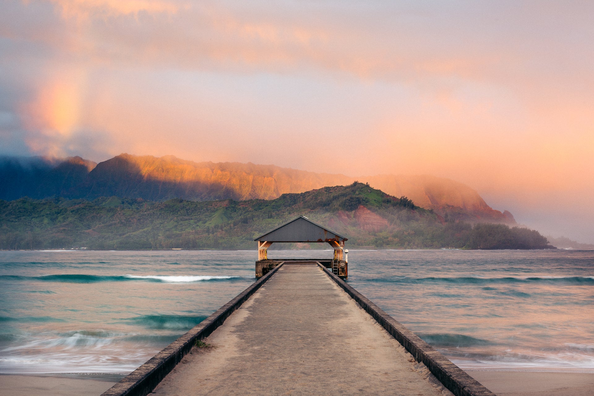 Long wooden pier extending into the ocean with a mountainous landscape and colorful sky.
